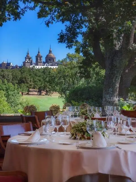 Outdoor terrace table set with wine glasses and white linens, with a monastery and gardens visible in the background