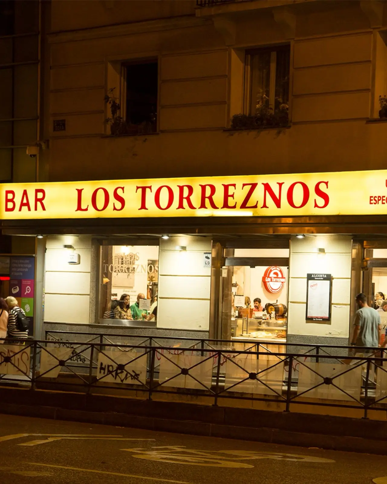 Night-time exterior storefront with an illuminated yellow sign reading BAR LOS TORREZNOS above lit windows showing patrons at the bar