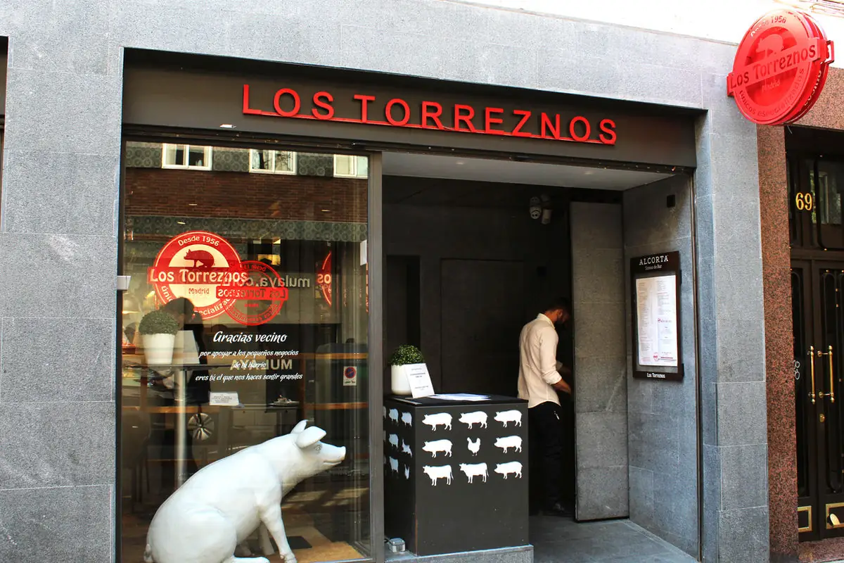 Daytime grey-stone storefront with red LOS TORREZNOS sign, a white pig statue in the window and a staff member in the doorway