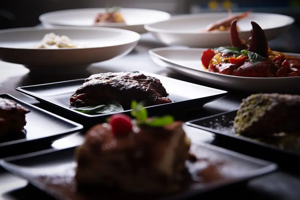 Table spread of dishes including a seared meat cut, tomato pasta with shellfish claws, a creamy pasta, and a chocolate dessert with raspberry and mint garnish, L'Osteria del Contadino