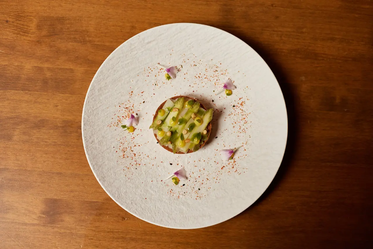 Round tart topped with green and white asparagus strips, citrus dots, and edible flowers on a textured white plate, L'Ós Panda