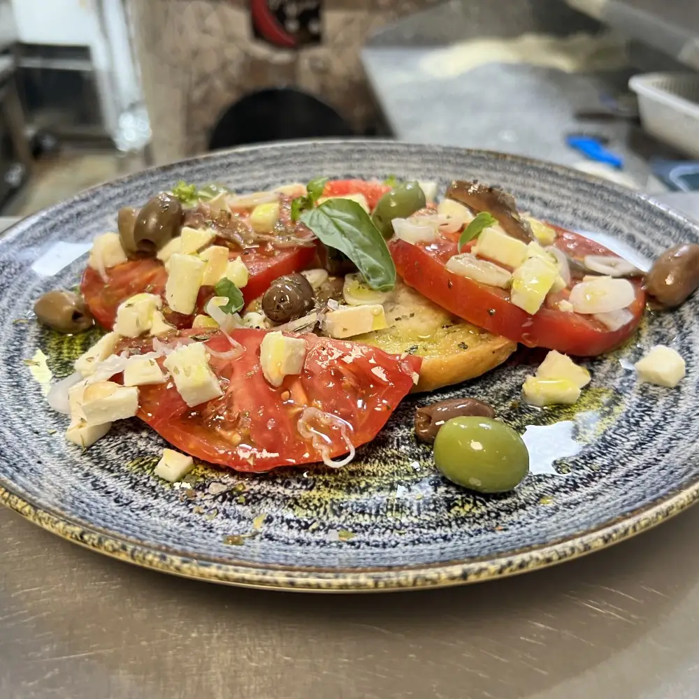 Sliced tomato bruschetta topped with cubes of cheese, green and black olives, capers and basil on a speckled blue ceramic plate