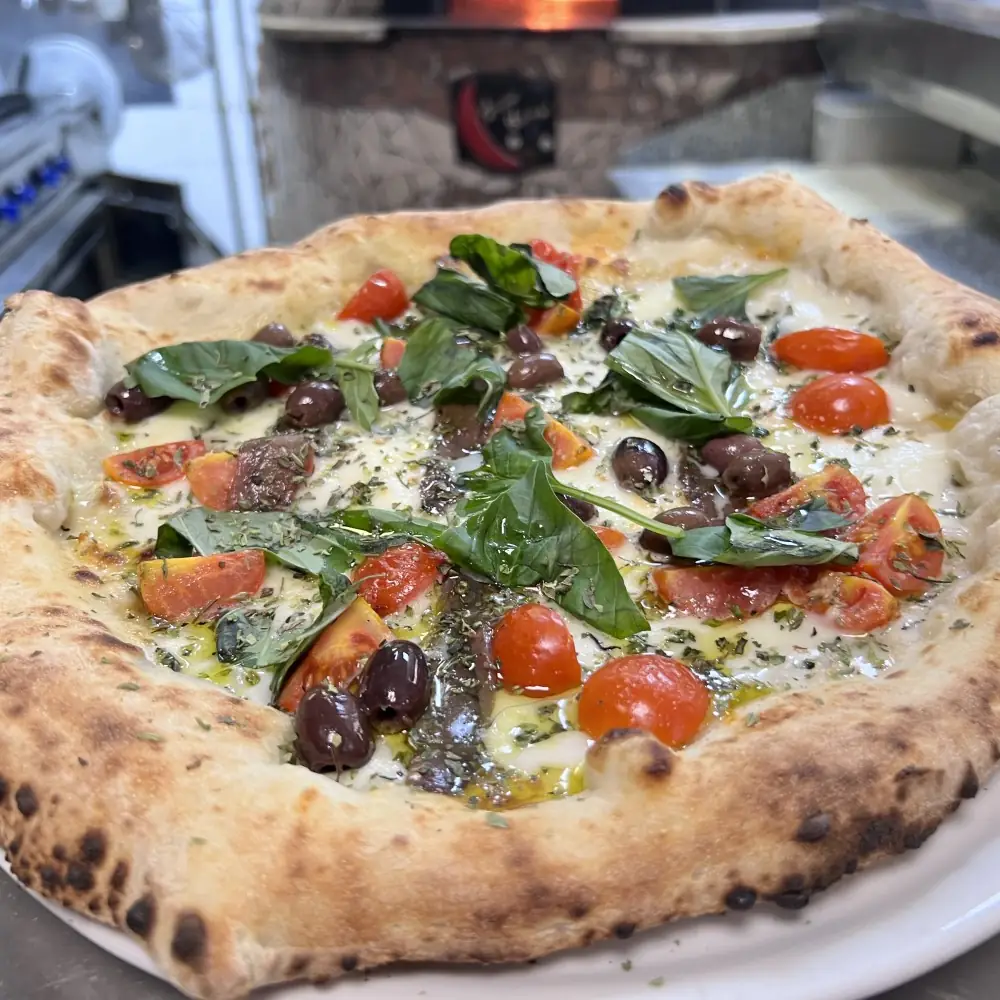 Wood-fired Neapolitan pizza with mozzarella, cherry tomatoes, black olives, oregano and fresh basil, puffed charred crust