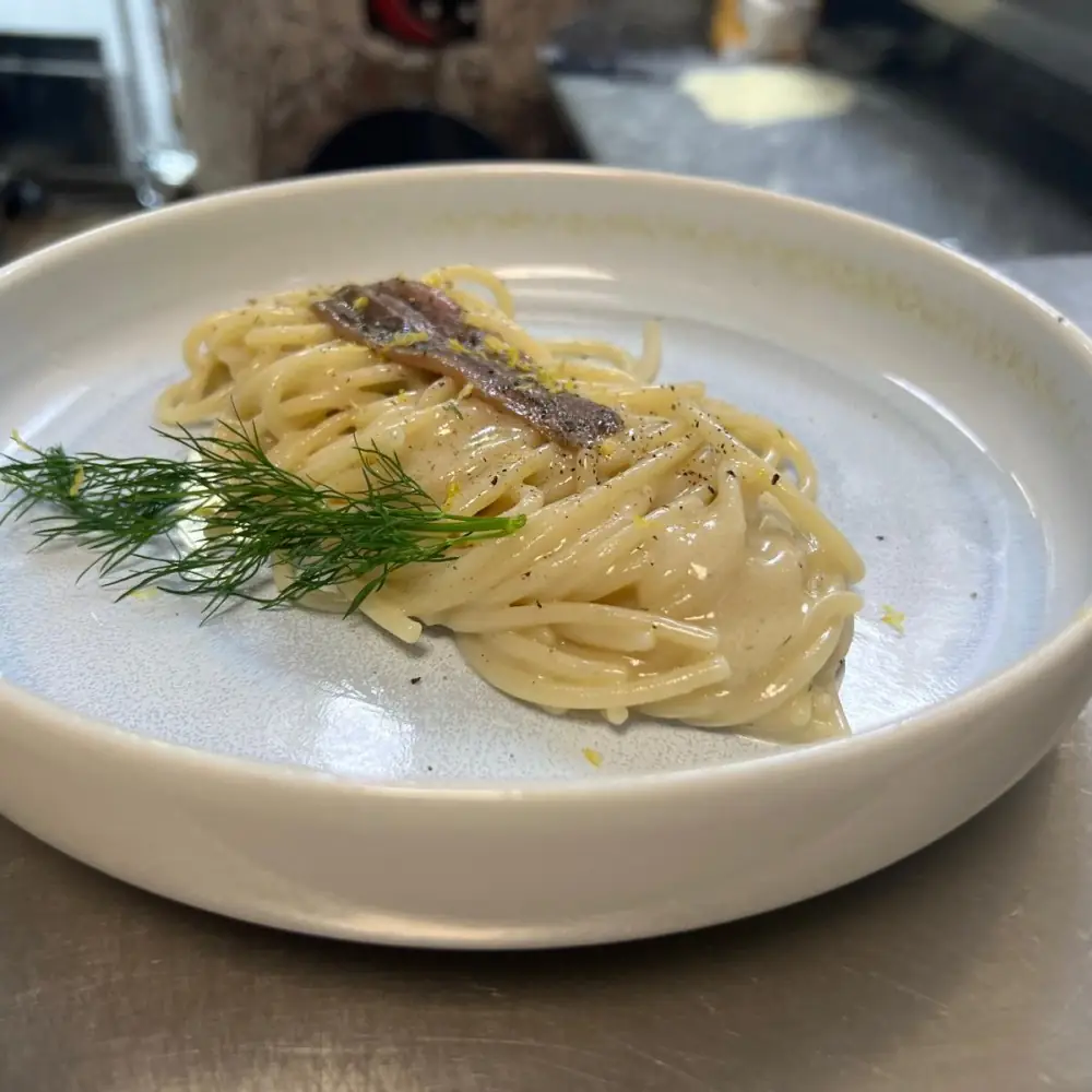 Nest of spaghetti in a creamy sauce topped with a whole anchovy fillet and a sprig of dill on a white plate