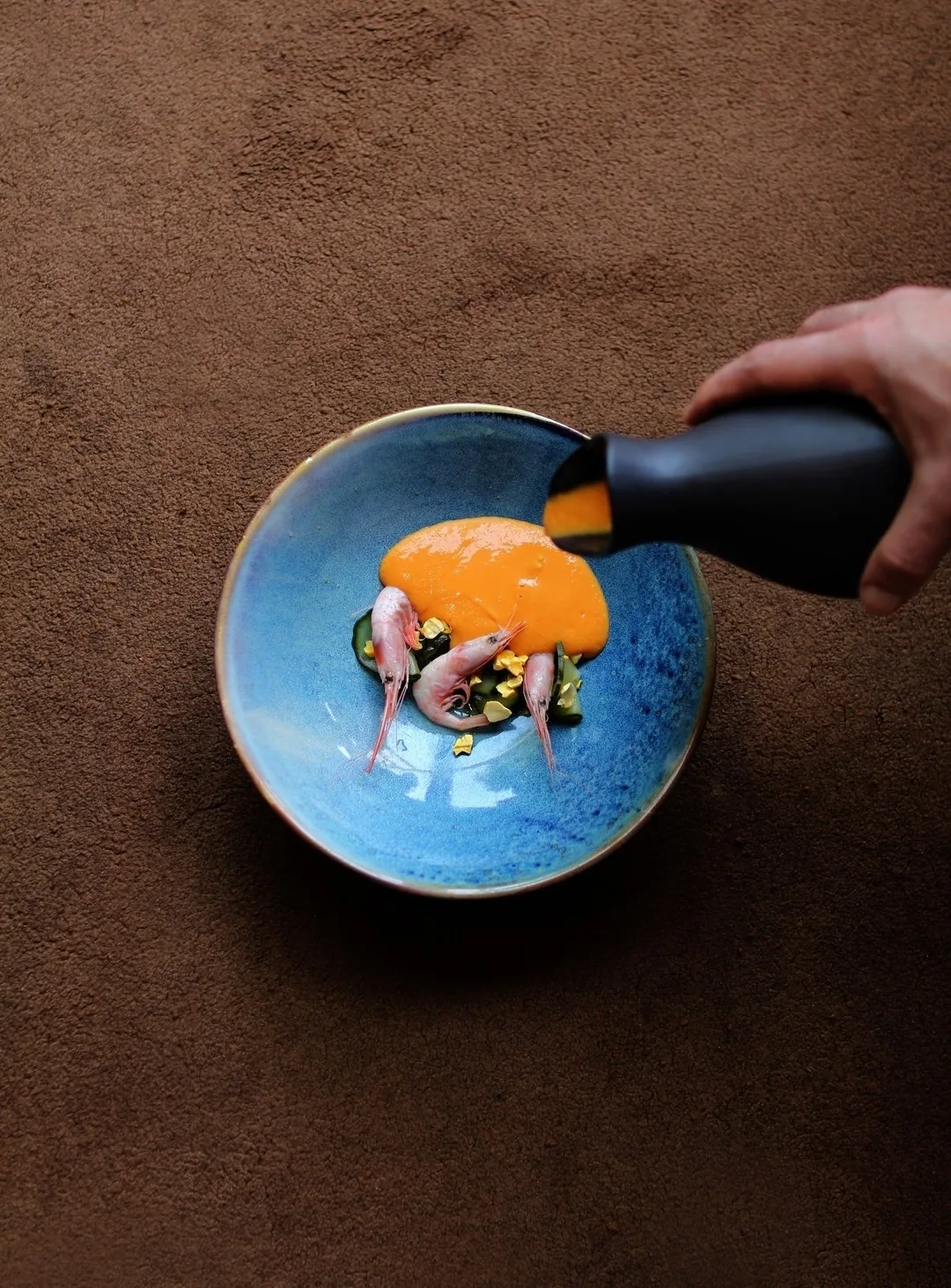 Small pink shrimp in a blue bowl with an orange sauce being poured over