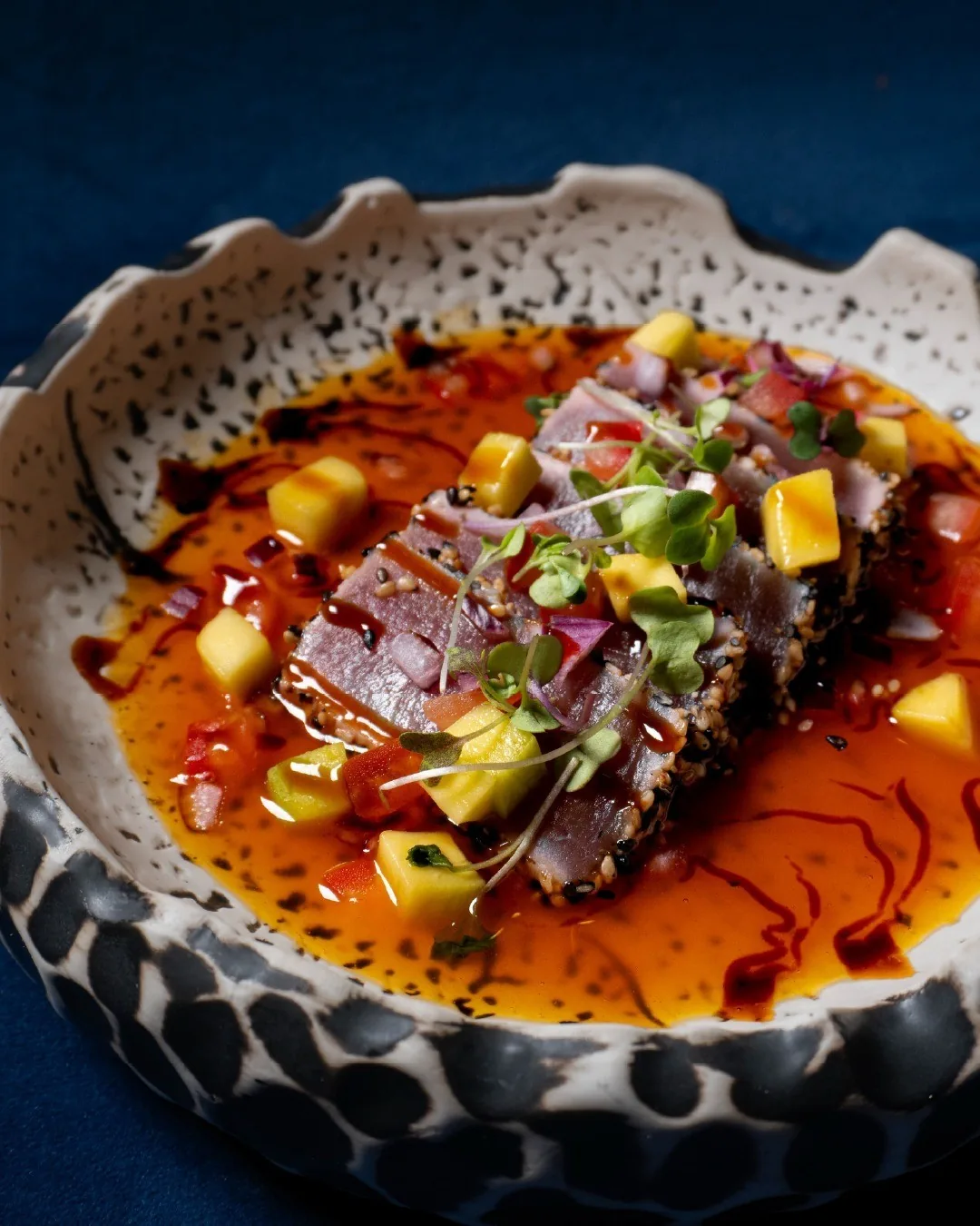 Slices of seared beef with diced mango and herbs in a red sauce in a dark speckled bowl