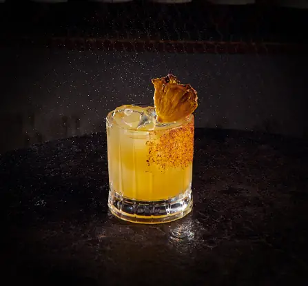 Yellow cocktail over ice in a rocks glass with a spiced rim and a dried pineapple slice garnish