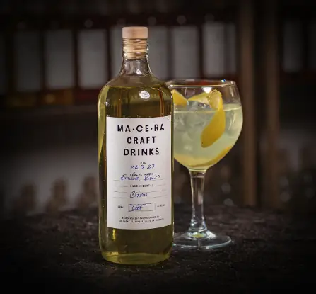 Macera Craft Drinks bottle next to a balloon glass drink over ice with a lemon peel twist