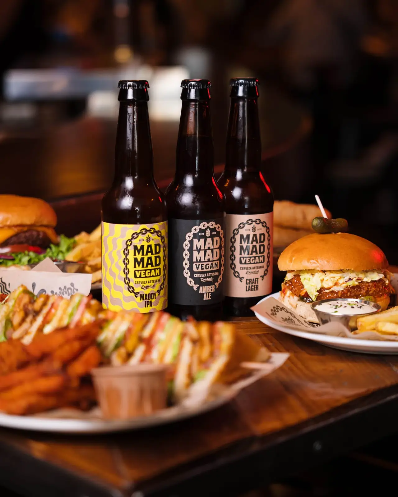 Two burgers on sesame buns, one topped with red cabbage slaw, next to golden fries and dipping sauces, with three craft beer bottles behind, at Mad Mad Vegan