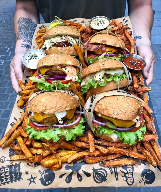 Overhead view of six sesame-bun burgers layered with lettuce, tomato, red onion, and melted cheese, surrounded by seasoned fries, at Mad Mad Vegan