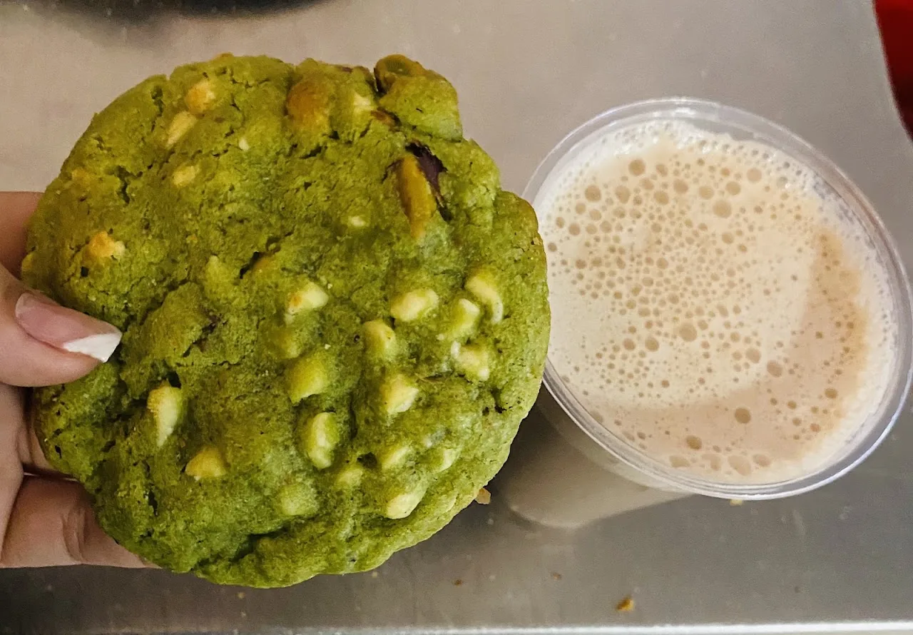 A pistachio cookie held next to an iced coffee drink