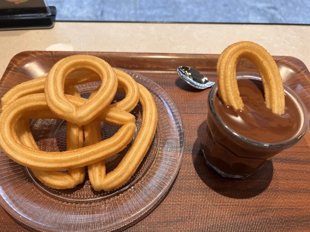 A plate of looped churros next to a glass of chocolate with a churro dipped in it, on a wooden tray