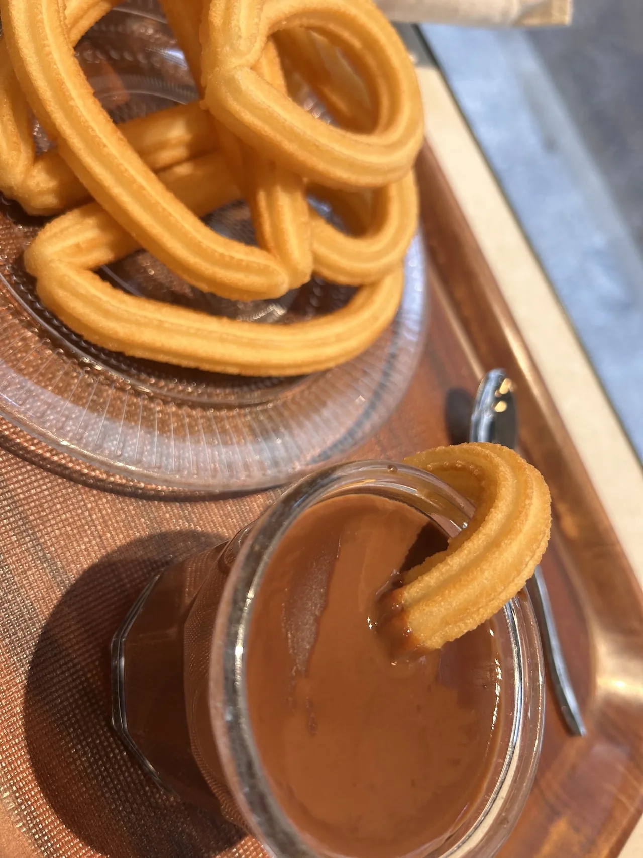 Looped churros on a glass plate above a glass of chocolate with a churro in it