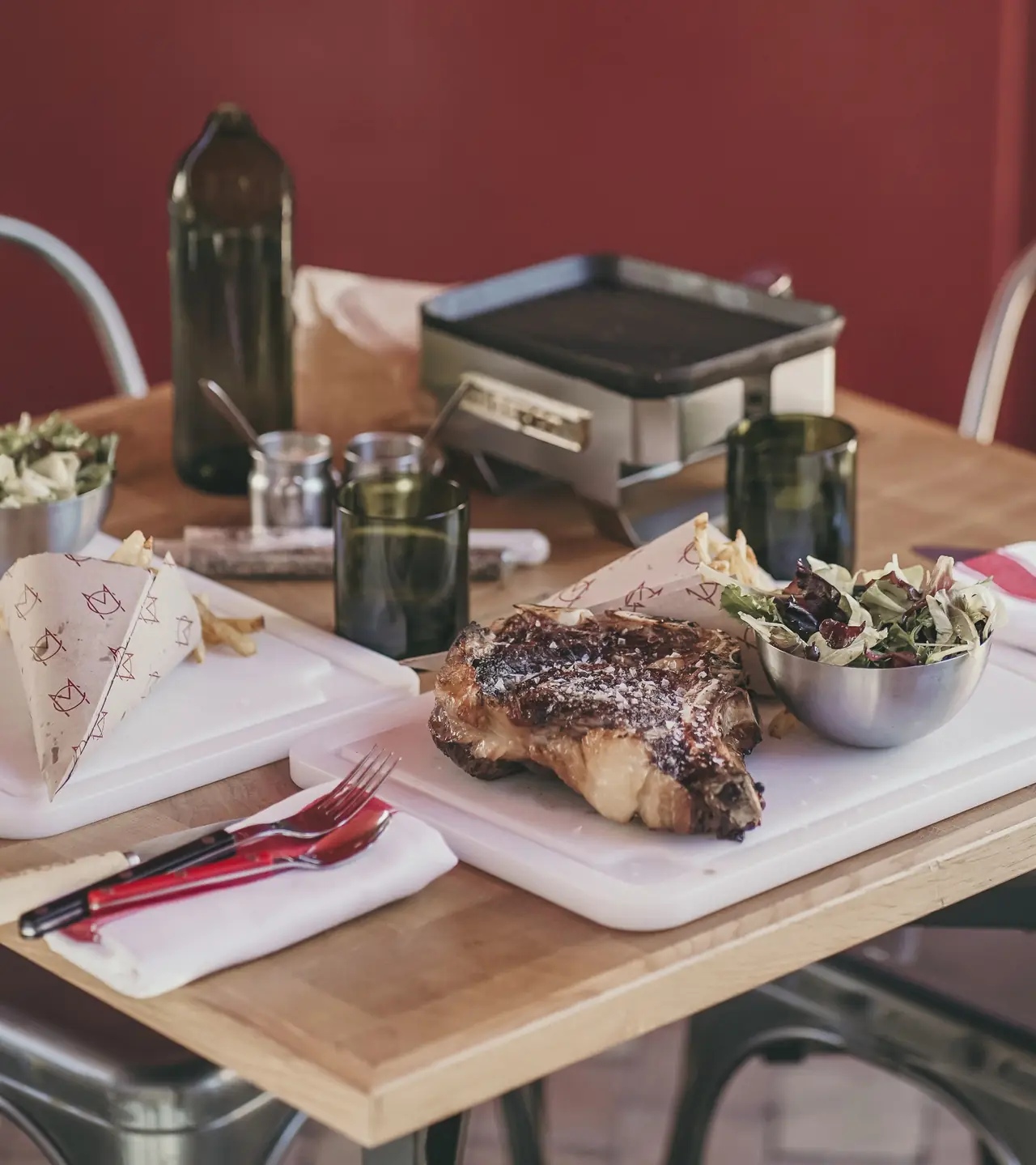 Grilled côte de boeuf with a side salad and a paper cone of fries on a wooden table, Maison Carne
