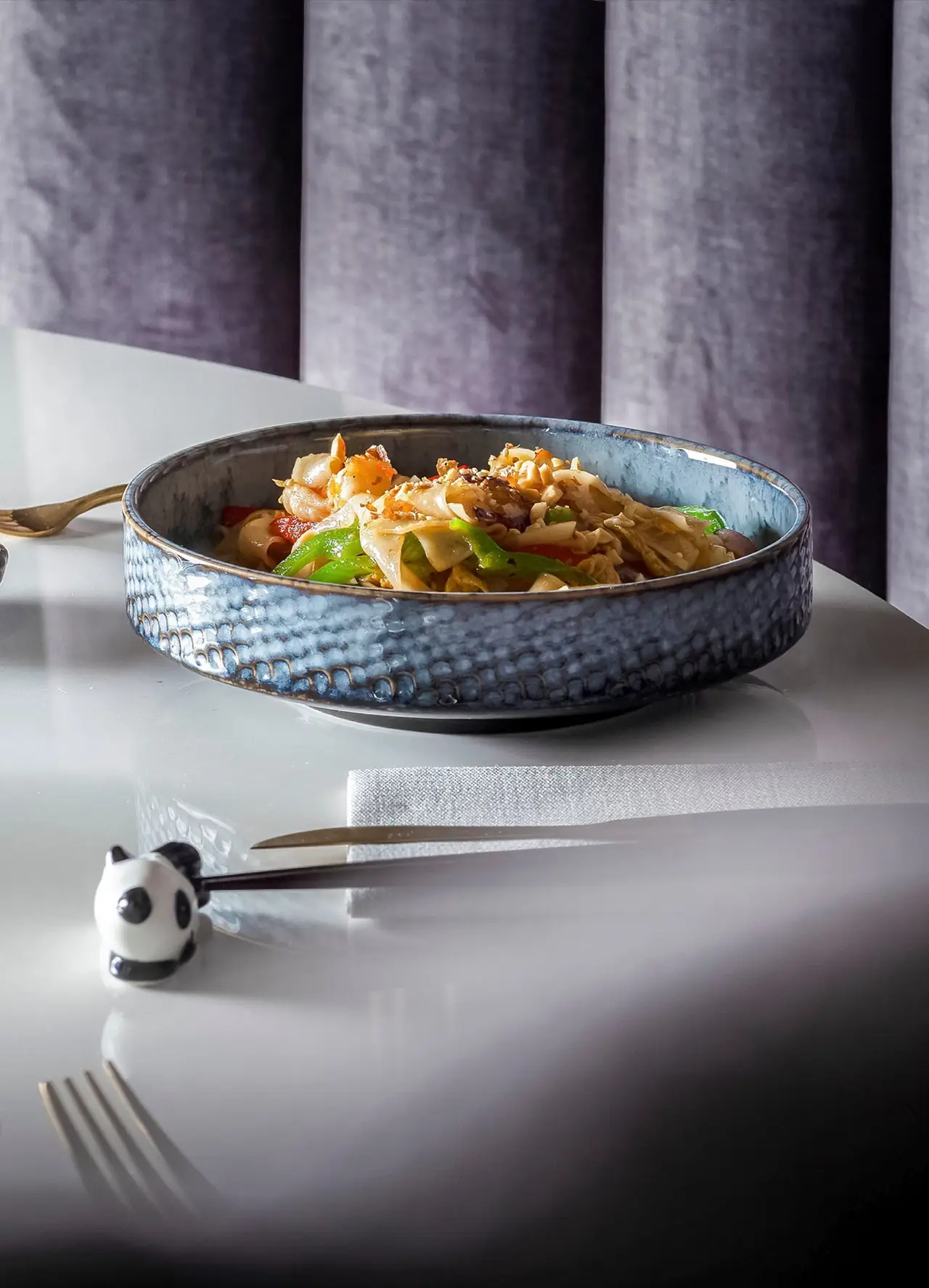 Stir-fried wide rice noodles with prawns, green pepper and peanuts in a blue ceramic bowl, panda chopstick rest alongside