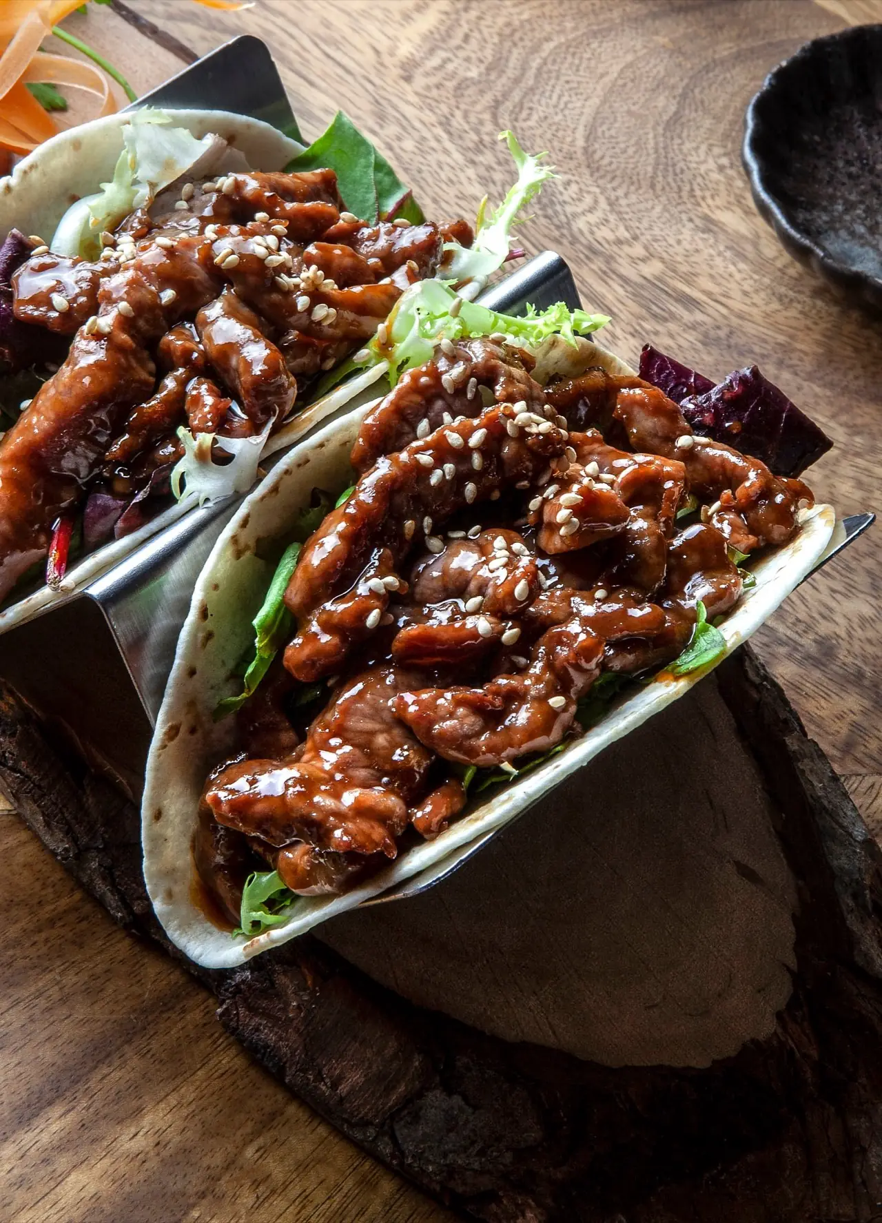 Two soft flour tacos filled with teriyaki-glazed meat strips, sesame seeds and salad leaves in a metal taco holder on a wooden board