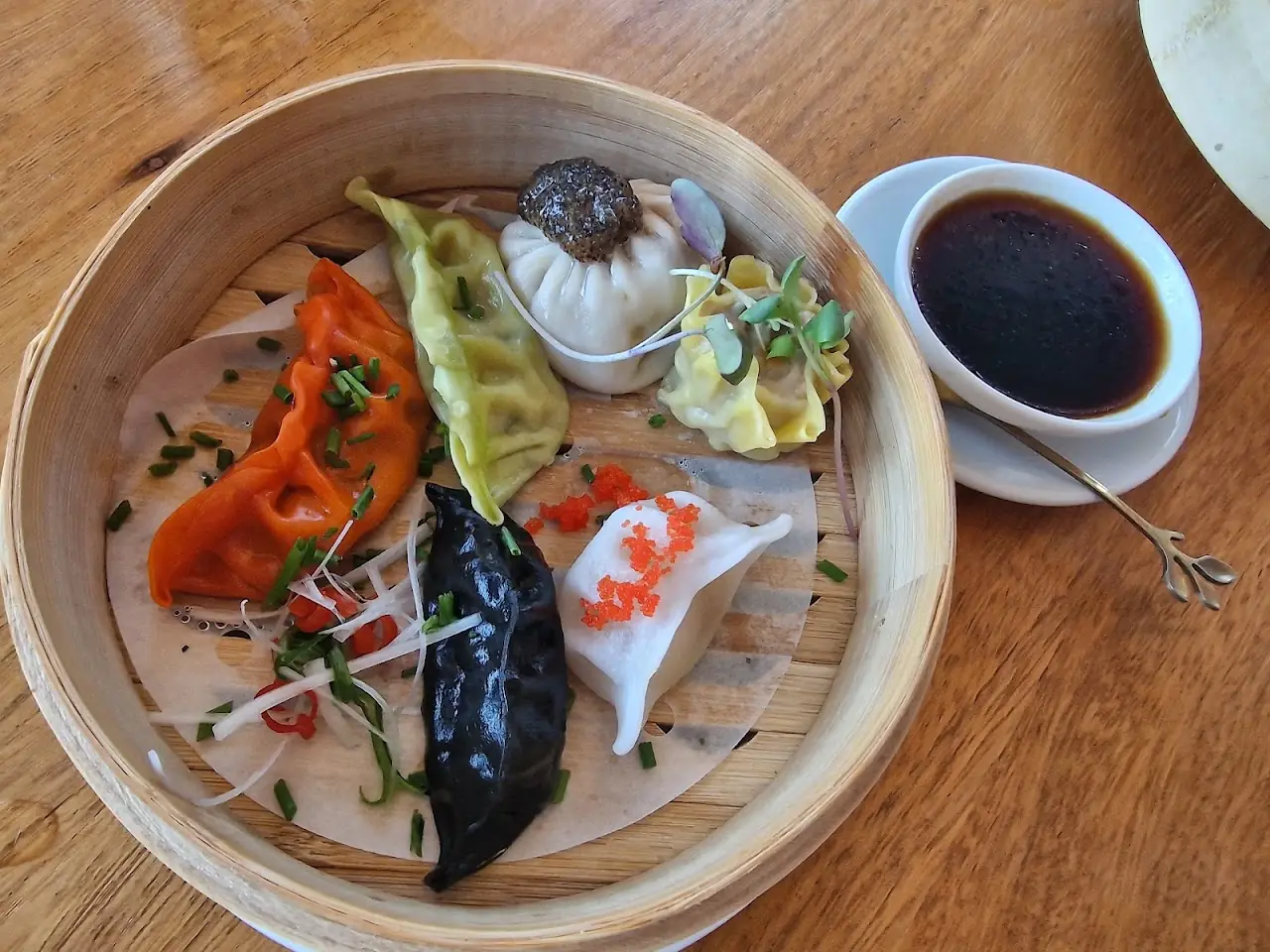 An assortment of colorful dumplings in a bamboo steamer with a dark dipping sauce