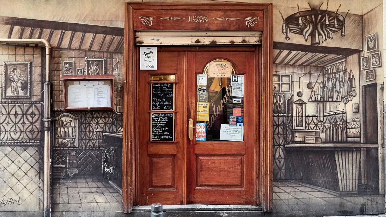 Malacatin storefront: a wooden double door dated 1895 set into a painted trompe-l'oeil mural of a tiled tavern, with handwritten chalkboard menu boards beside it