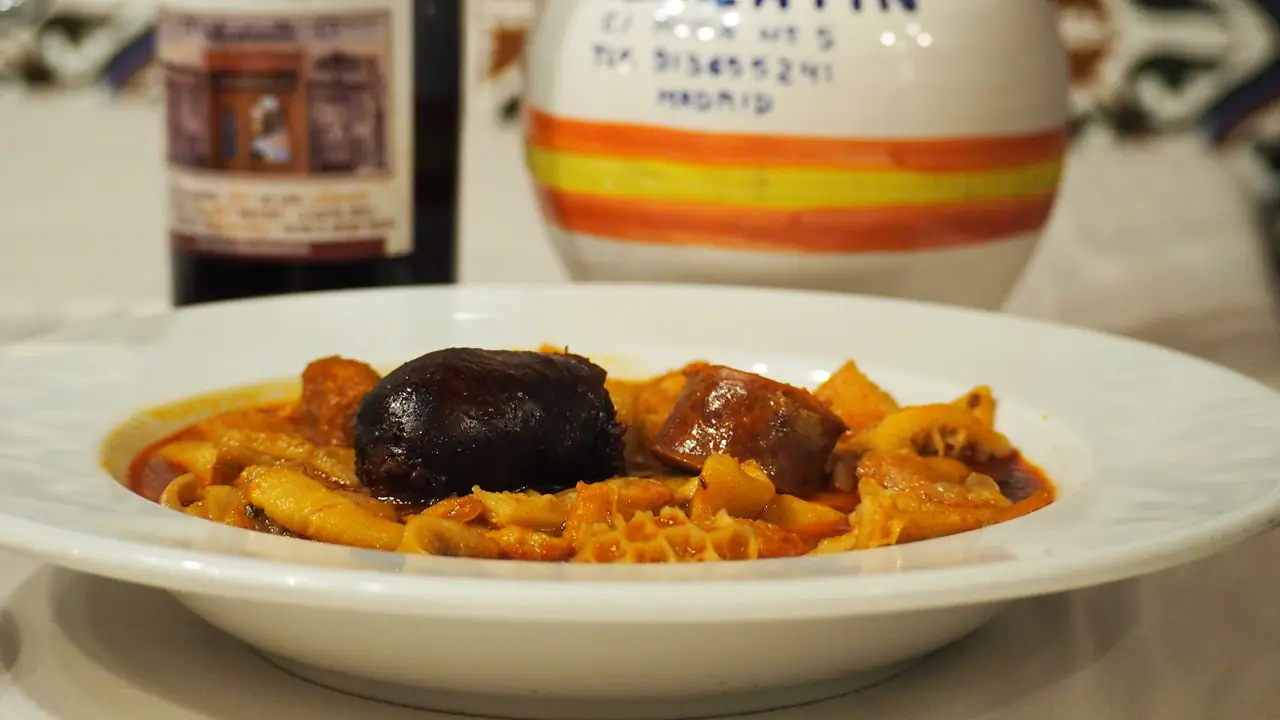 Bowl of callos madrileno-style stew with pasta, blood sausage and sliced chorizo in red tomato broth, a labelled jug behind