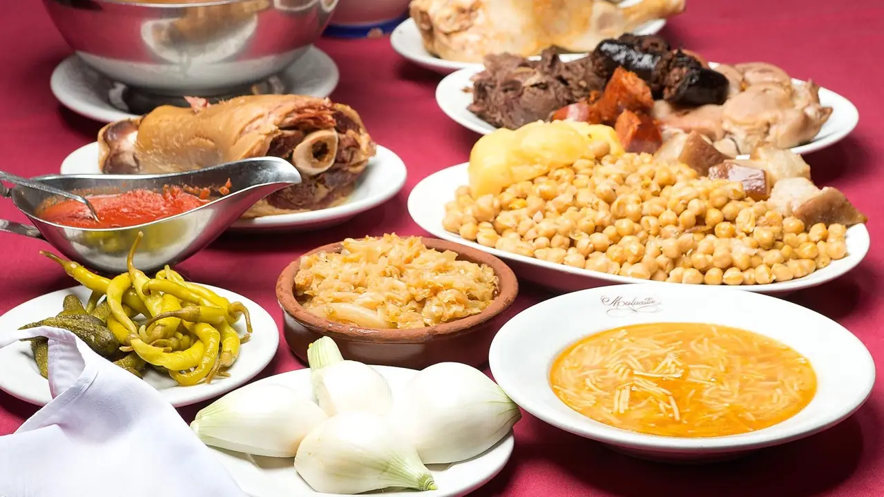 Full cocido madrileno spread on a red tablecloth: plates of chickpeas with pork and vegetables, boiled meats and ham hock, pickled peppers, cabbage, onions and a bowl of noodle soup