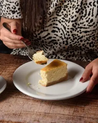 Slice of baked cheesecake on a white plate, a diner lifting a forkful, hands and torso behind