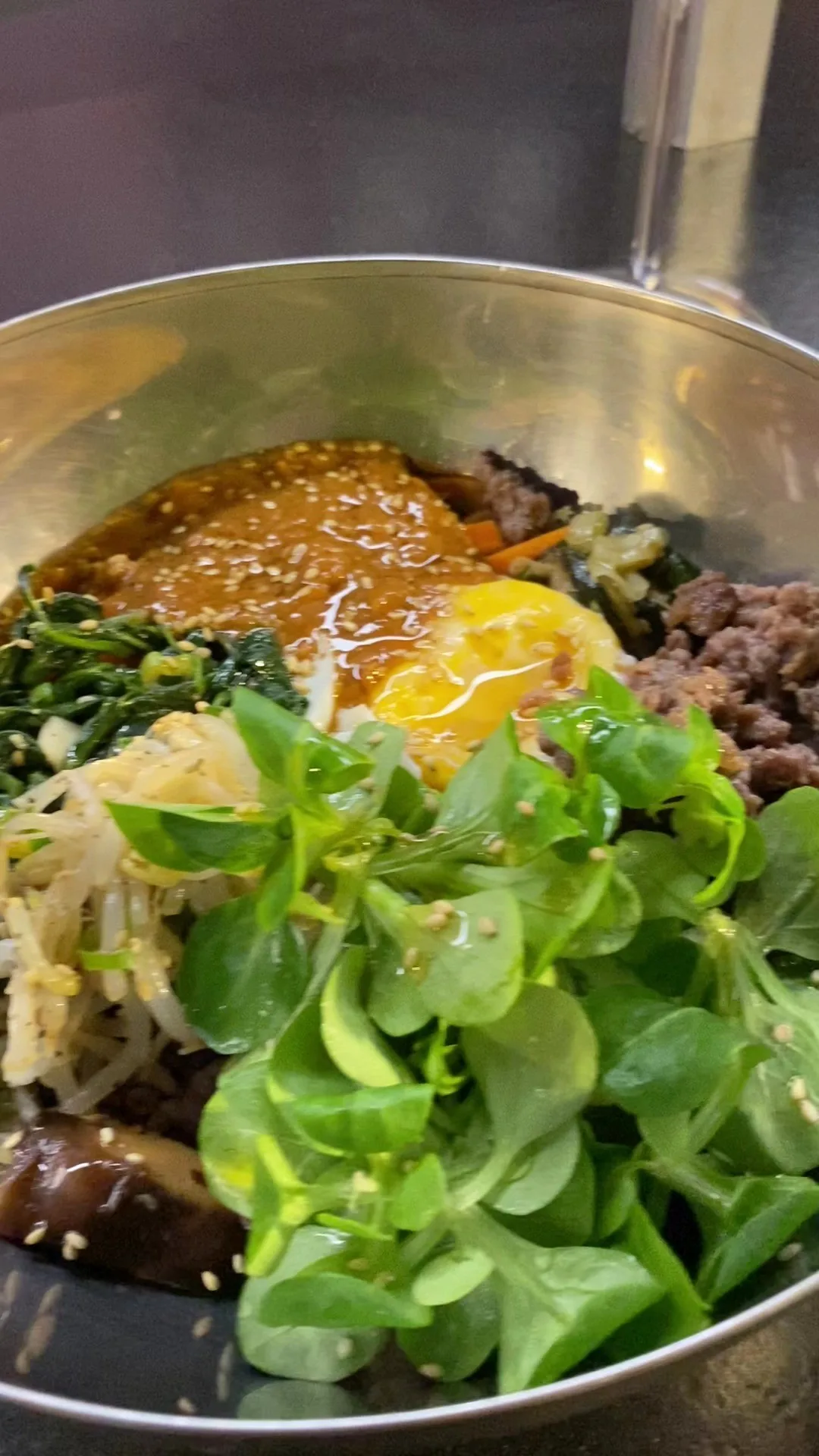 A metal bowl of rice topped with fresh greens, sauce and sliced ingredients