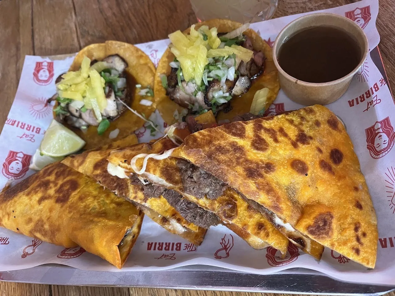Tacos and a crisp folded quesadilla with a small cup of dipping broth on a paper-lined tray
