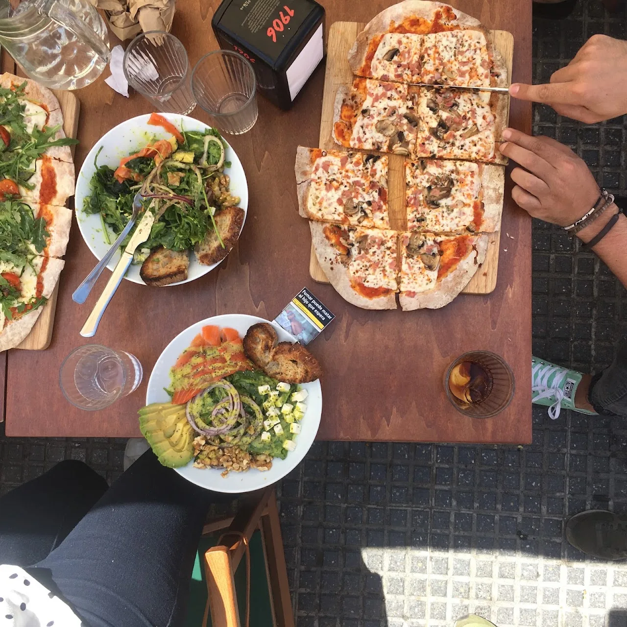 An overhead view of a wooden board pizza and salad bowls shared on a table