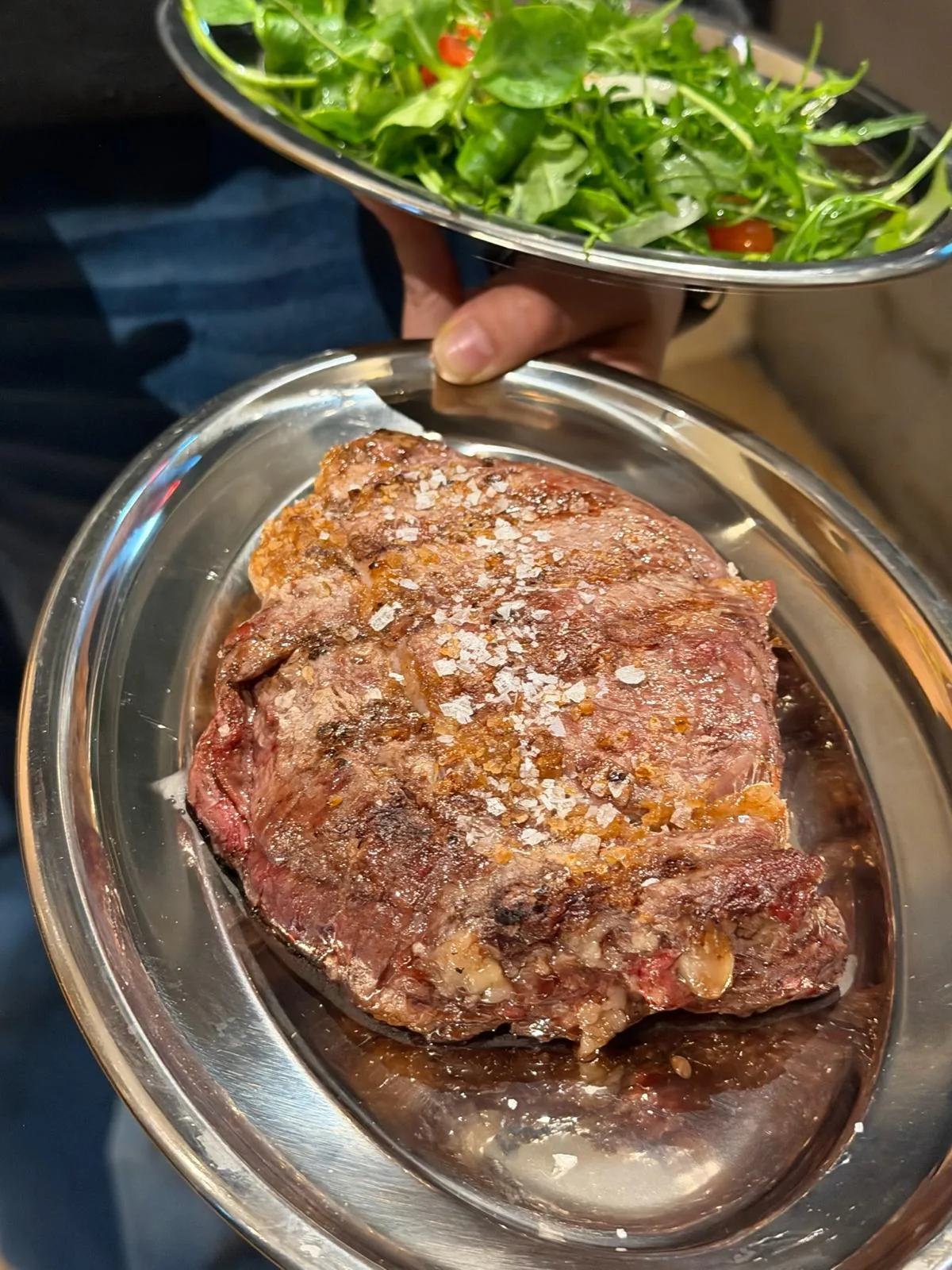 A hand holding a green salad bowl above a plate of grilled steak