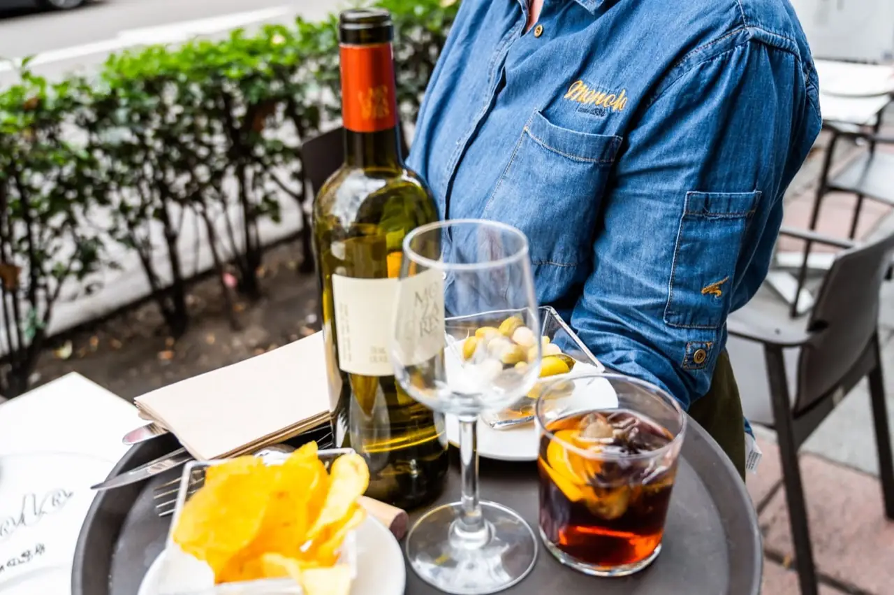 Cafe table on the terrace with a bottle of white wine, an empty glass, a vermut over ice with orange, a dish of olives and a bowl of potato chips; a waiter in a denim shirt stands behind