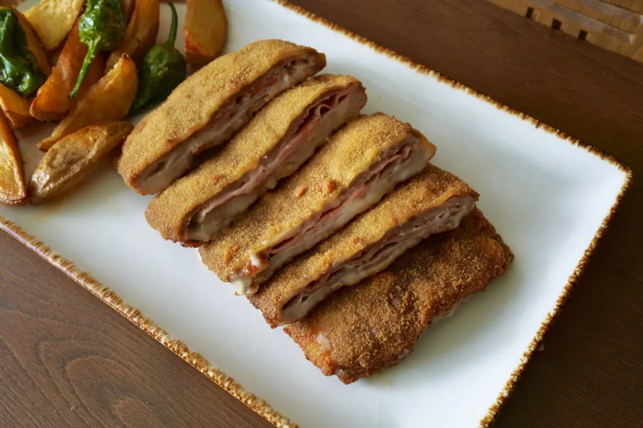 Sliced breaded cachopo filled with ham and cheese on a white platter, with fried potato wedges and padron peppers