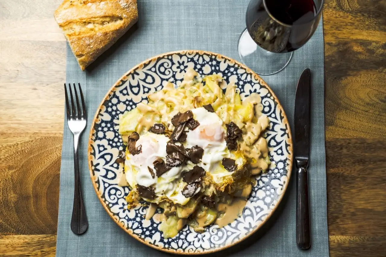 Broken fried eggs over potatoes with cream sauce and black truffle shavings on a blue-patterned plate, bread and a glass of red wine alongside