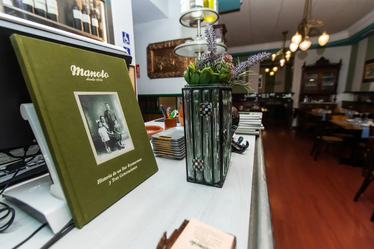 Interior looking down the dining room over the counter, a green 'Manolo desde 1934' history book propped up, set tables with wood chairs beyond