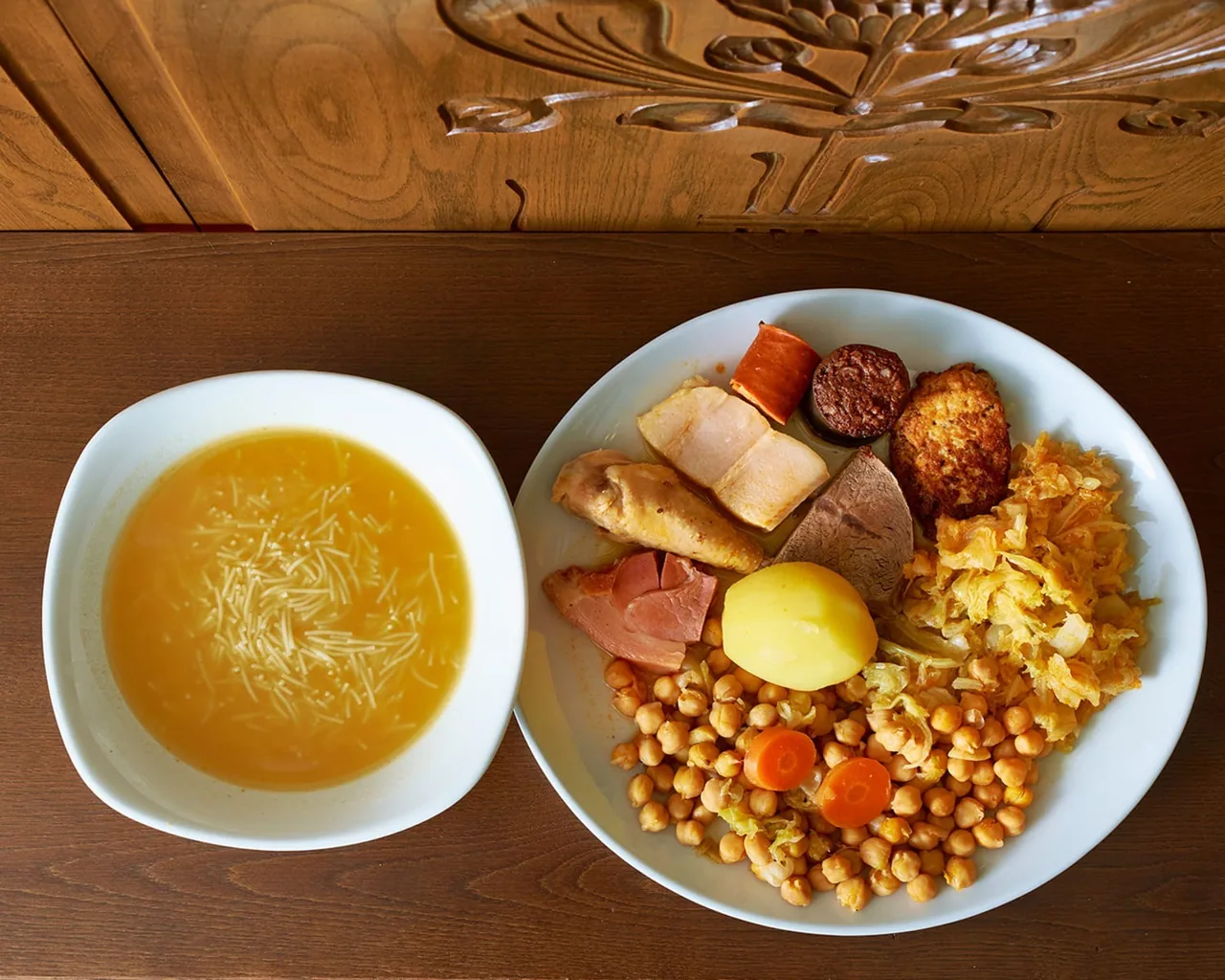 A white bowl of noodle broth beside a plate of chickpeas, potato, carrot, chorizo, black pudding and boiled meats on a wooden table
