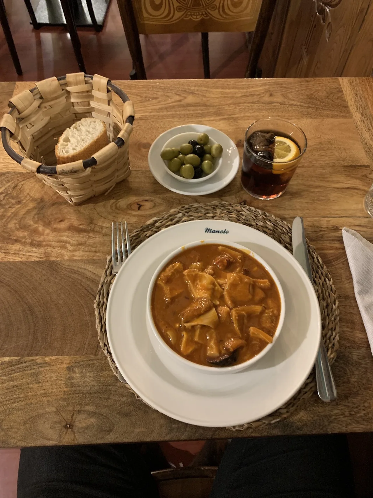 A bowl of tripe stew on a plate marked Manolo, with a basket of bread, olives and a glass of vermouth
