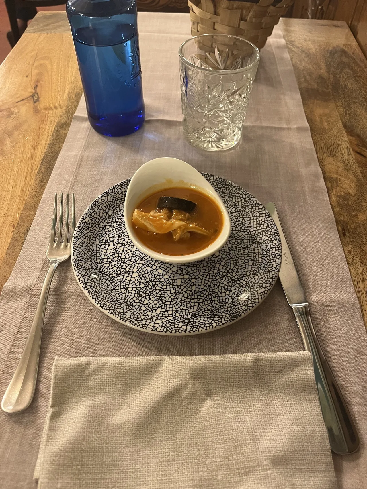A small white bowl of tripe stew on a patterned plate beside a fork, knife and blue water bottle