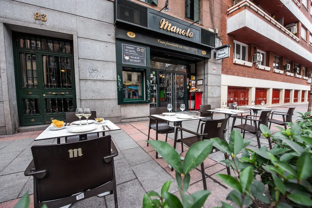 A green and dark wood restaurant frontage with a sign and tables set outside on the pavement.
