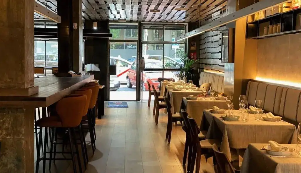 Interior of a narrow trattoria dining room with a wooden bar and orange stools on the left, banquette seating and tables set with white linens and wine glasses on the right, street visible through the front glass door
