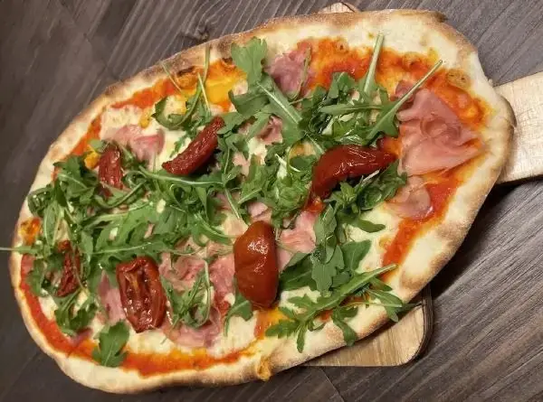 Oval thin-crust pizza topped with cooked ham, arugula and semi-dried tomatoes on a wooden board over a dark table