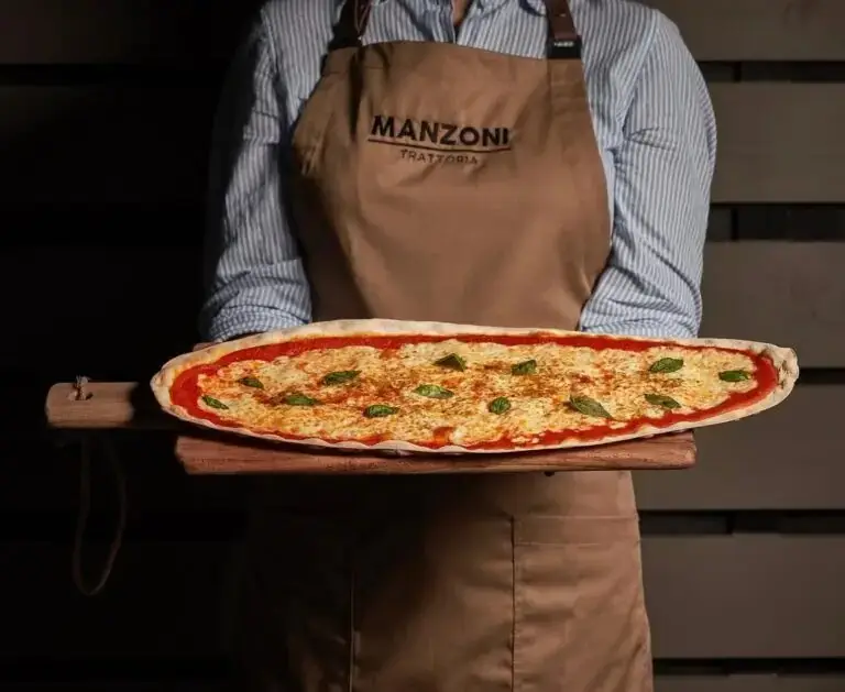Long oval margherita pizza with tomato, melted cheese and basil on a wooden peel, held by a person in a striped shirt and branded apron