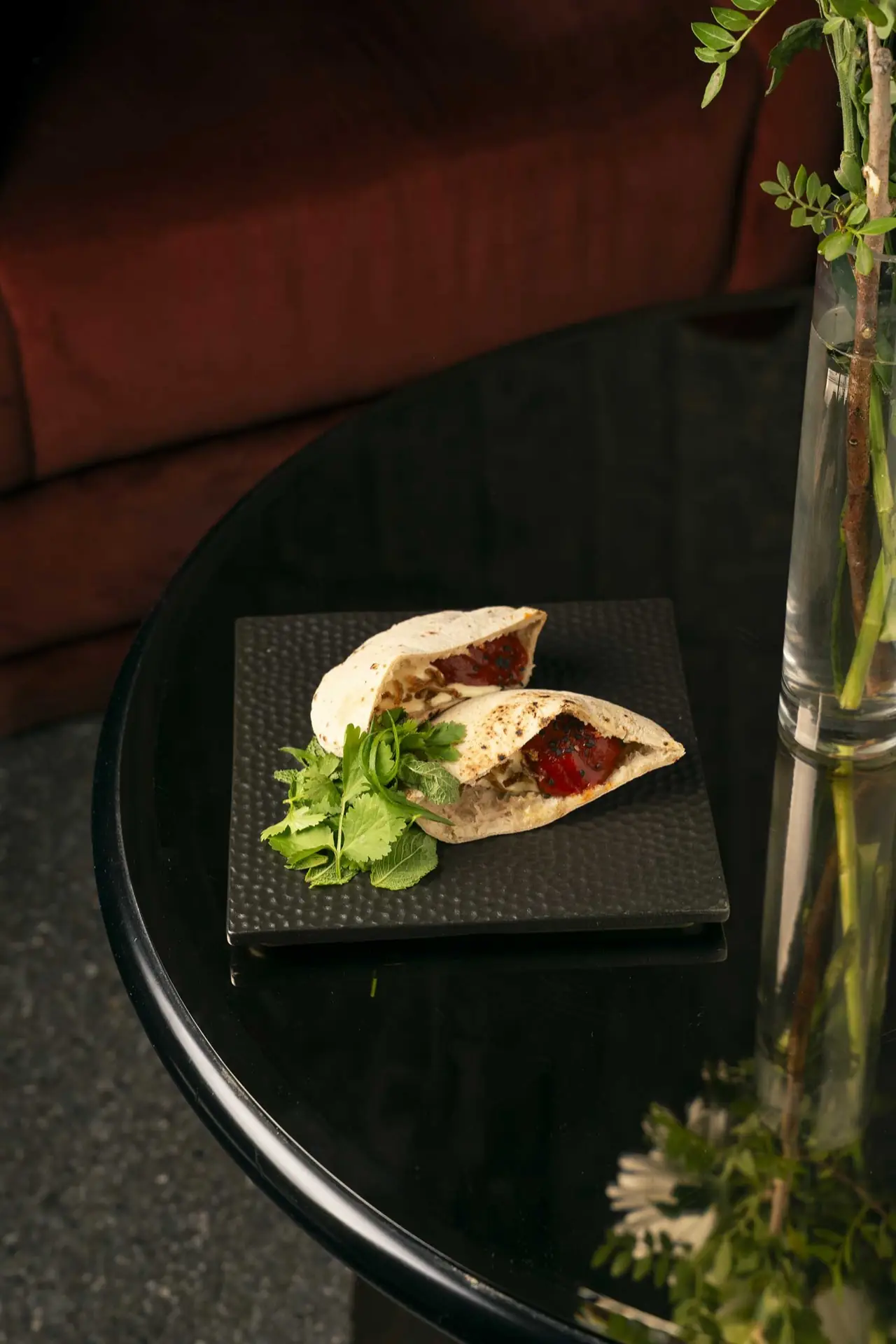 Halved stuffed pita filled with roasted red pepper and a dark sauce, with a bunch of fresh coriander and mint on a black square plate on a glossy black table