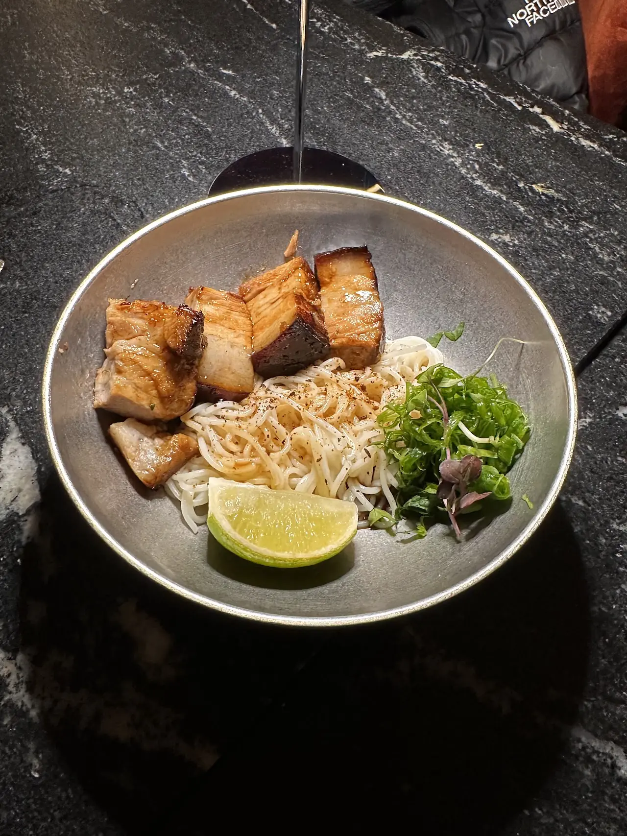 Seared pork belly pieces over rice noodles with herbs and a lime wedge