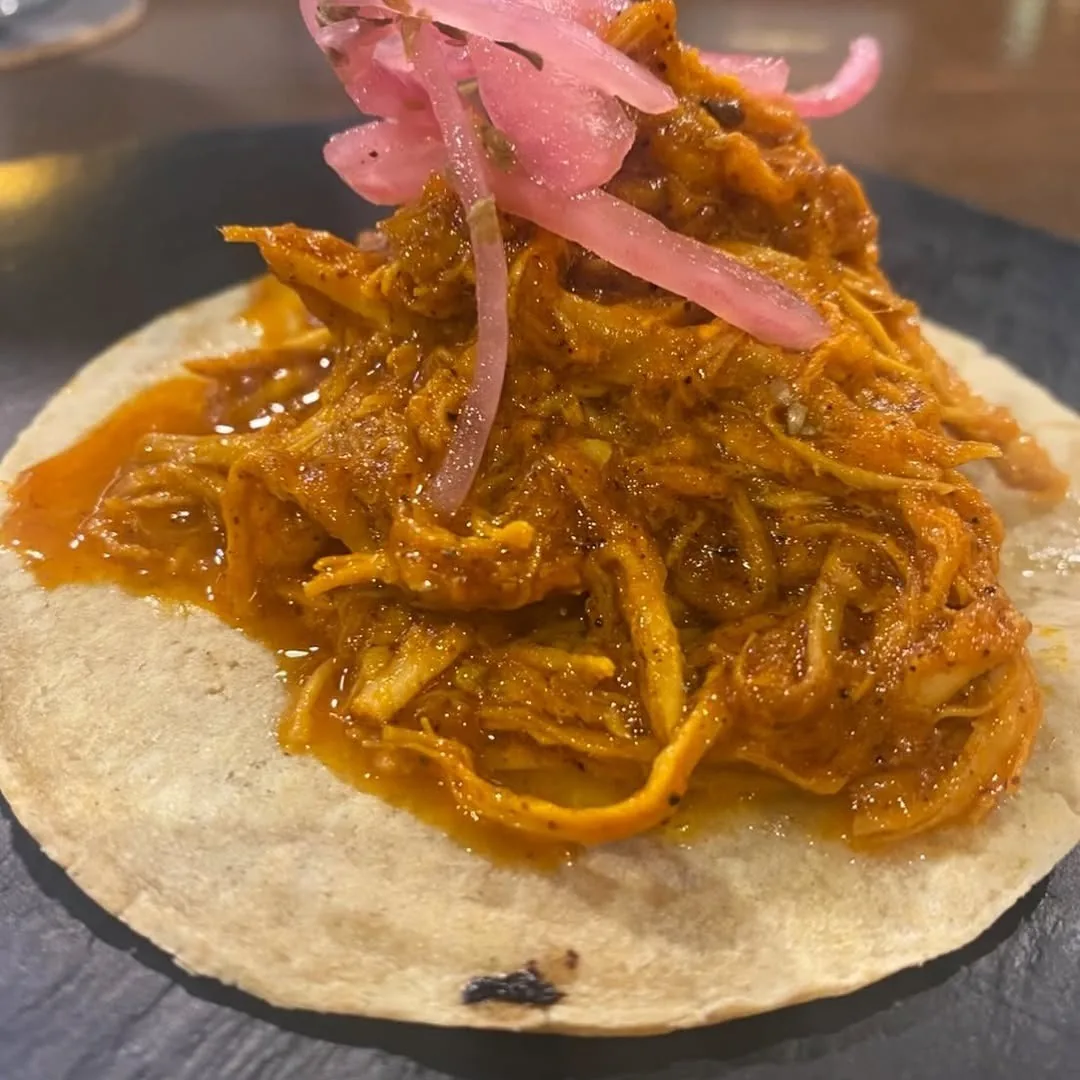 A tortilla topped with shredded orange meat and pink pickled onion on a dark plate