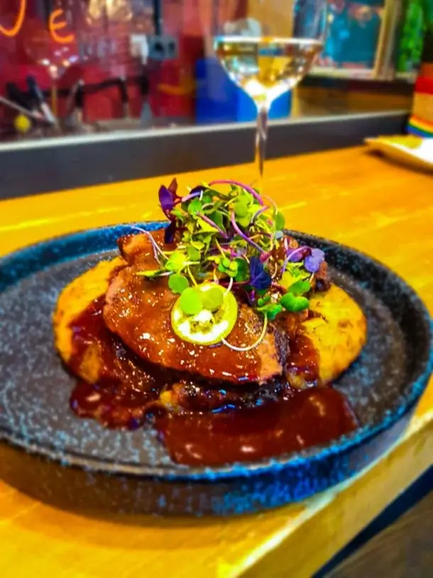 Glazed braised meat over parmentier with red sauce and a tuft of microgreens on a blue stoneware plate, a glass of white wine behind