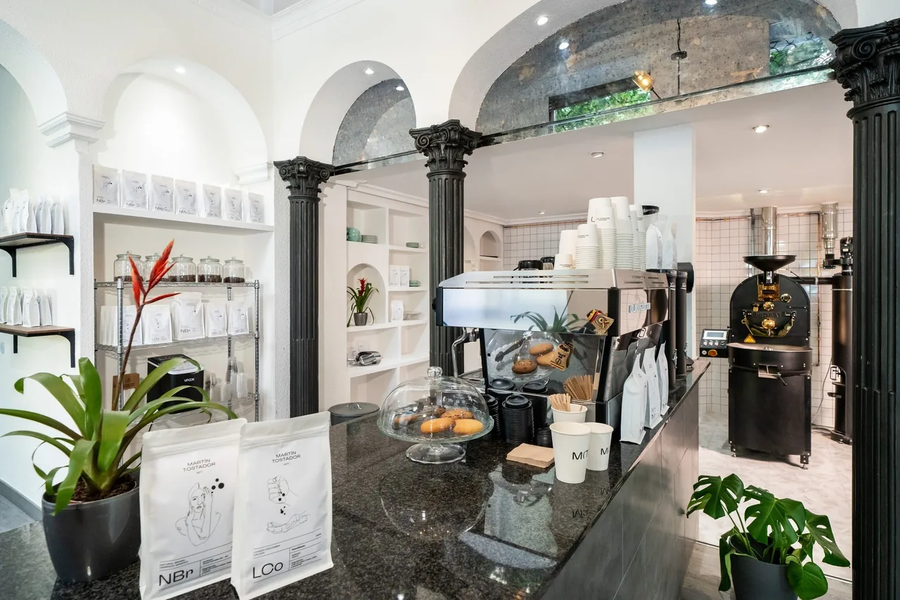 A bright white cafe with black columns, an espresso machine on a granite counter, bags of coffee and a roasting machine in the back room