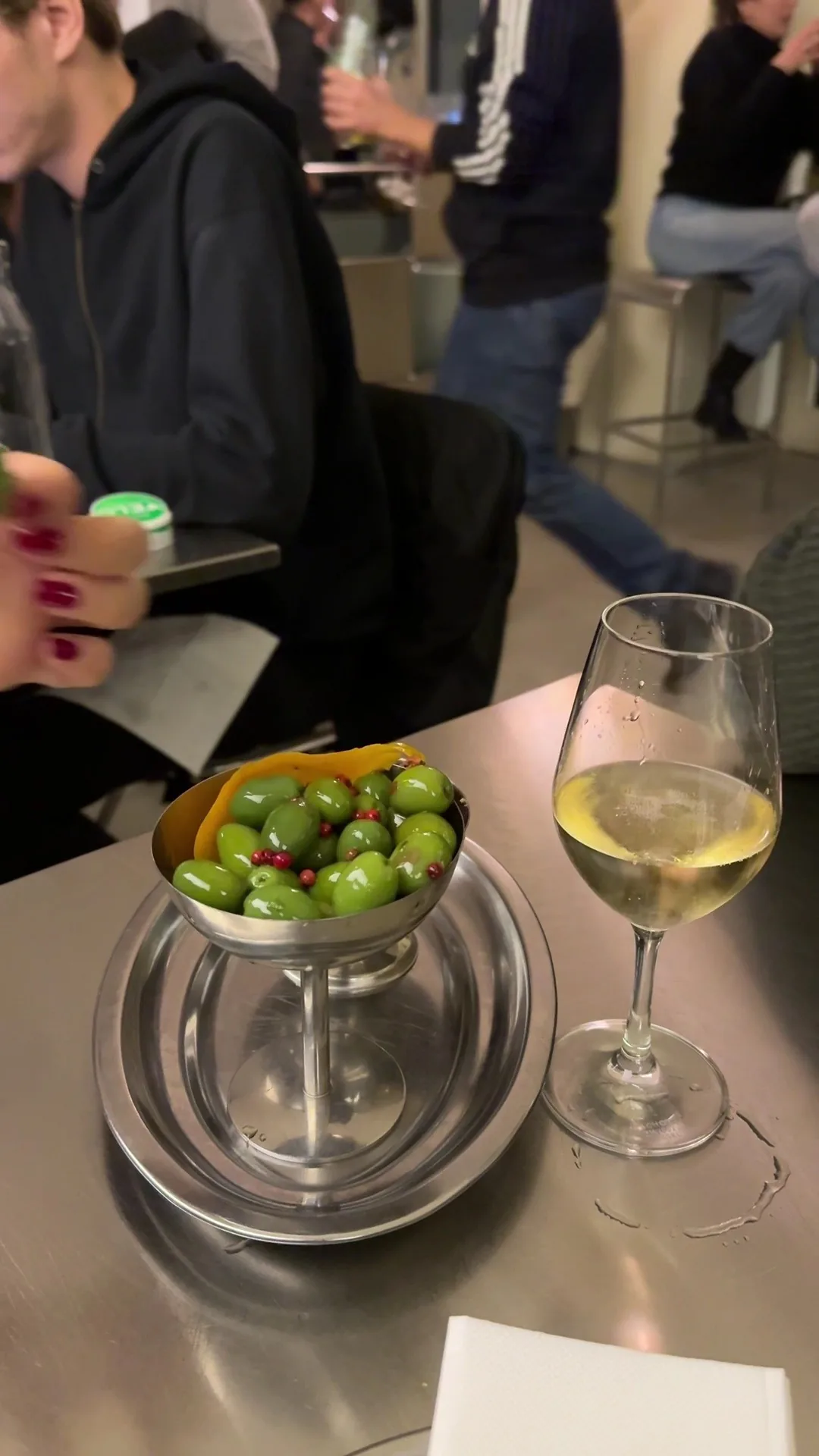 A glass of white wine and a small bowl of green olives on a steel counter