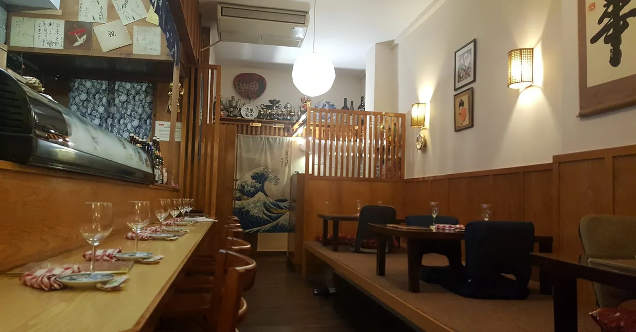 Japanese dining room with a wooden sushi counter and refrigerated display case on the left, places laid with wine glasses and chopsticks, and a raised tatami area with low tables and floor cushions beyond a Hokusai wave curtain