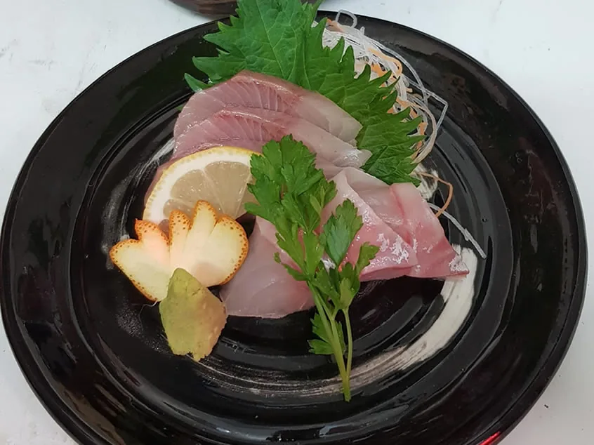 Slices of pale pink sashimi fanned on a black lacquer plate with a shiso leaf, shredded daikon, a lemon slice, parsley and a mound of wasabi