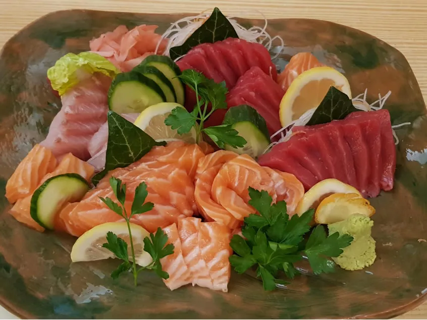 Green ceramic platter of mixed sashimi - deep red tuna, orange salmon and pale white fish - with cucumber slices, lemon wedges, parsley, shredded daikon, pickled ginger and wasabi