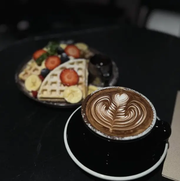 Latte with rosetta latte art in a black cup and saucer beside a plate of waffles with strawberries, banana and blueberries